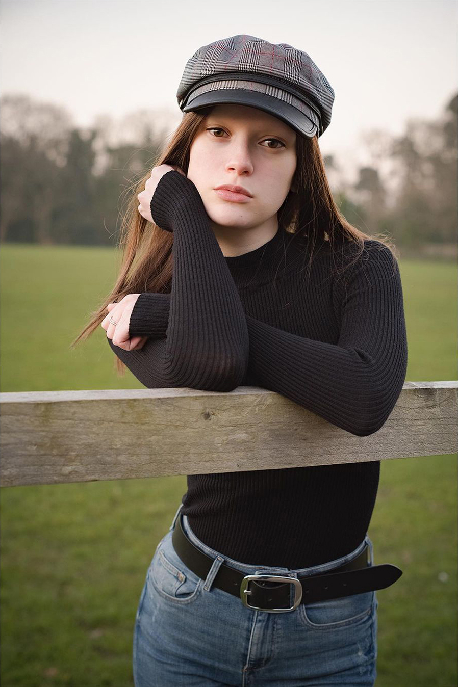 Joelle wearing a wig and hat while leaning on a fence.