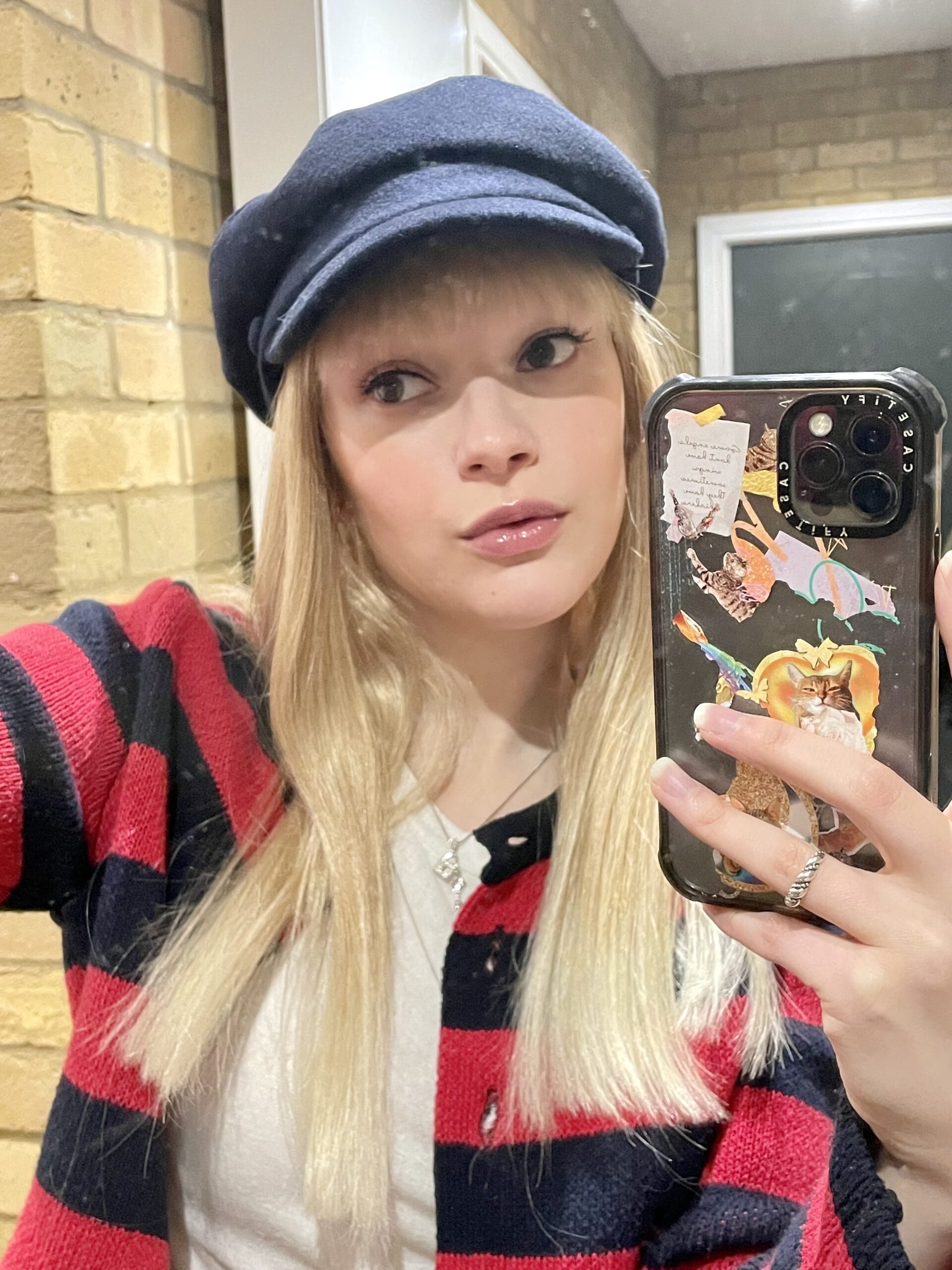 A picture of actress and singer Joelle taking a selfie in a blonde wig wearing a navy bake boy hat and a red and navy striped cardigan.