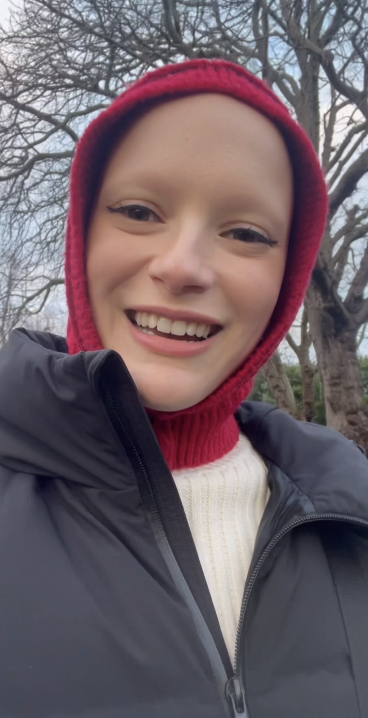 A picture of actress and singer Joelle smiling dressed in a black puffer coat, white jumper and a knitted red hood walking outside.